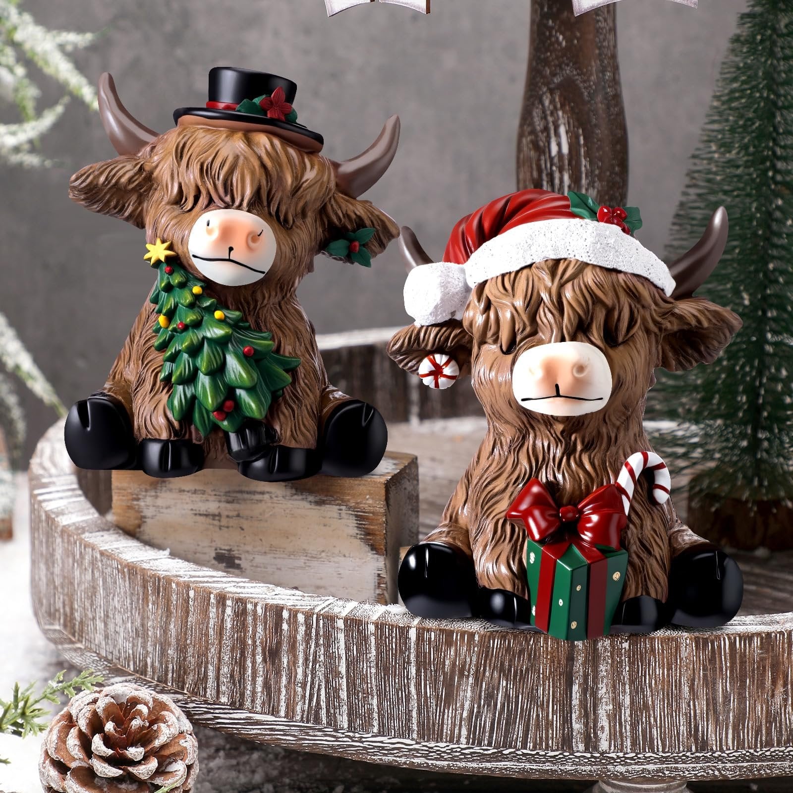 Two resin highland cow figurines in festive holiday attire, one with a Christmas tree and top hat, the other with a Santa hat and gift.