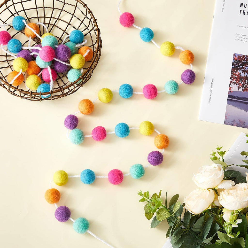 Colorful wool felt ball garland draped from a basket, with decorative flowers and a magazine in the background.