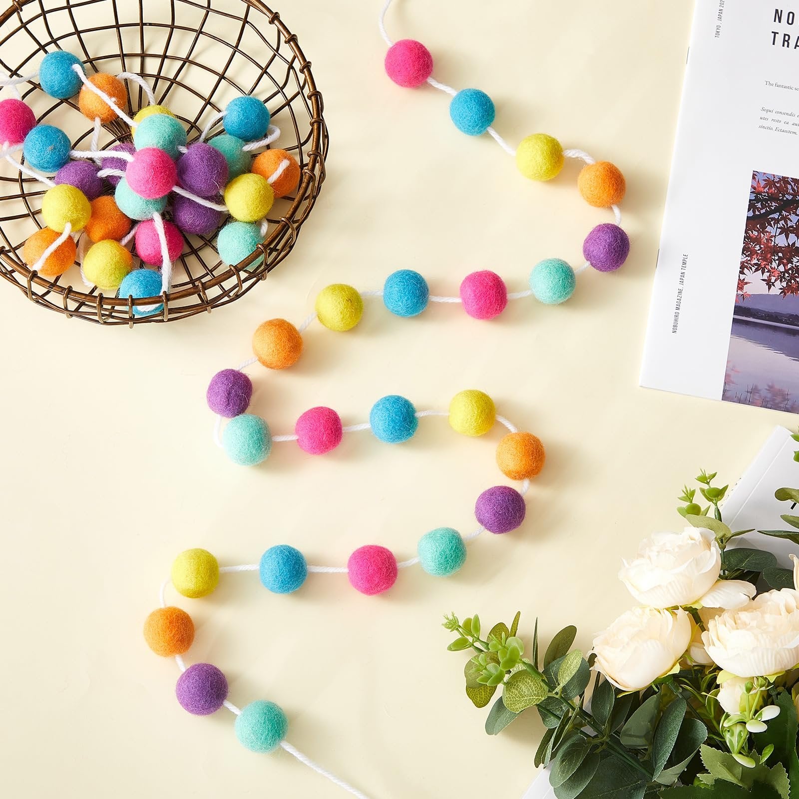 Colorful wool felt ball garland draped from a basket, with decorative flowers and a magazine in the background.
