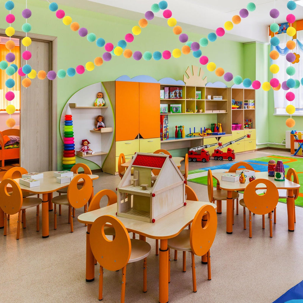 Colorful wool felt ball garlands strung across a classroom ceiling above tables and chairs.
