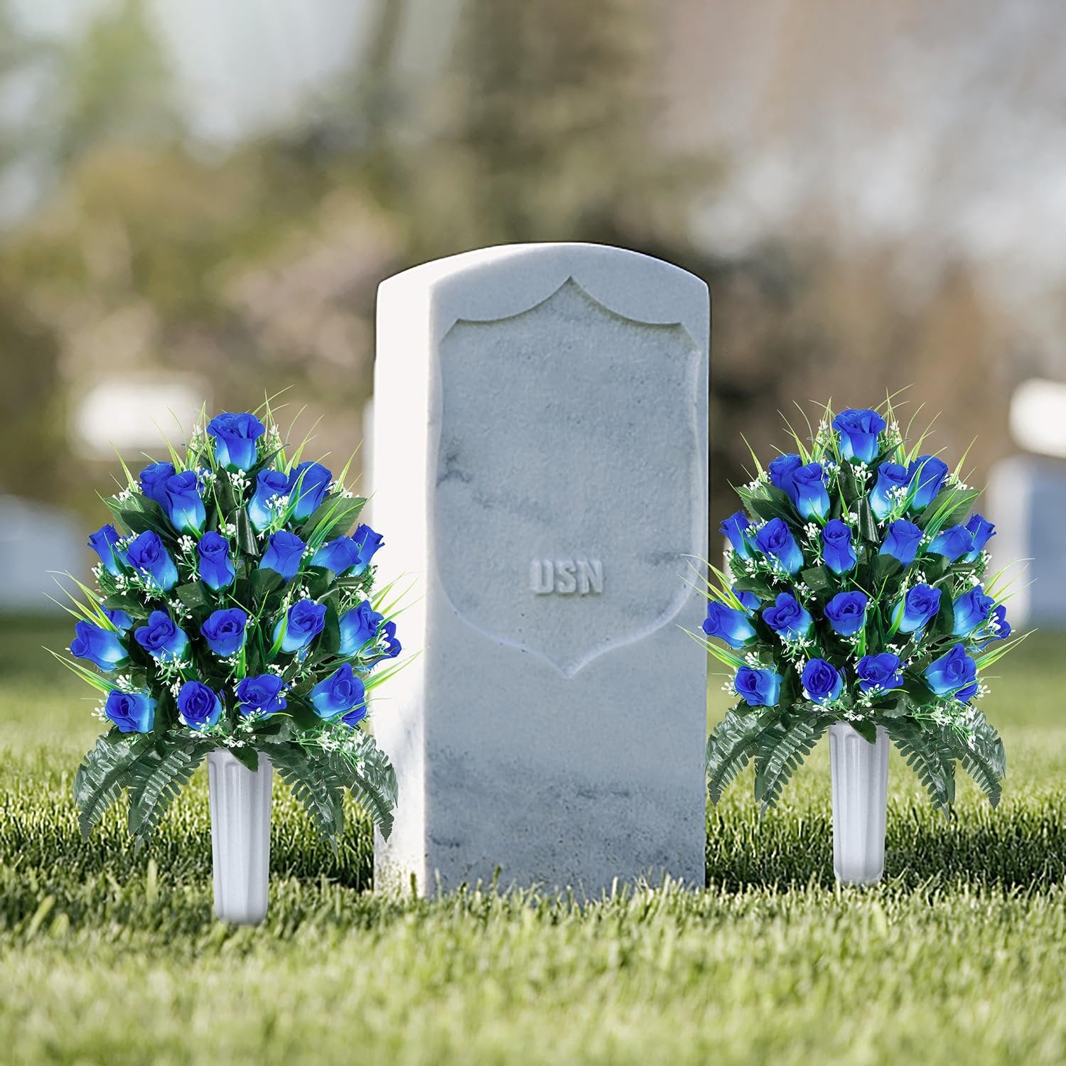 Two bouquets of blue artificial roses in white vases placed on grass in front of a tombstone with 'USN' inscription.
