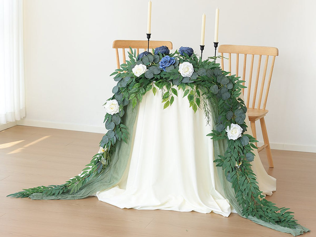 Artificial greenery garland with white and blue roses draped over a table between two chairs, with candles on the table.