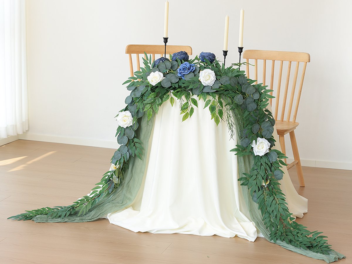 Artificial greenery garland with white and blue roses draped over a table between two chairs, with candles on the table.