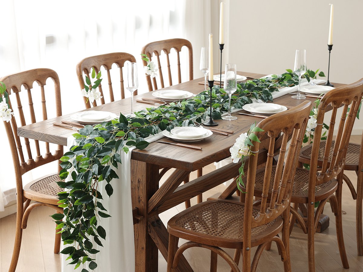 Dining table set for a meal with artificial greenery garland and white flowers on chairs