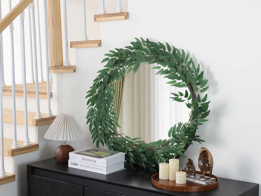 Artificial eucalyptus garland draped around a circular mirror on a dresser, with a lamp and books nearby.