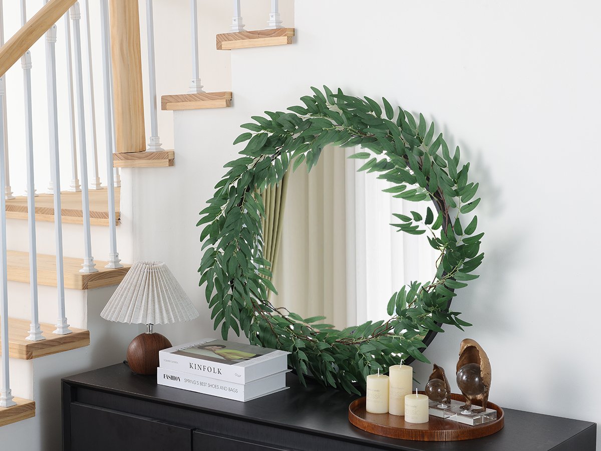 Artificial eucalyptus garland draped around a circular mirror on a dresser, with a lamp and books nearby.