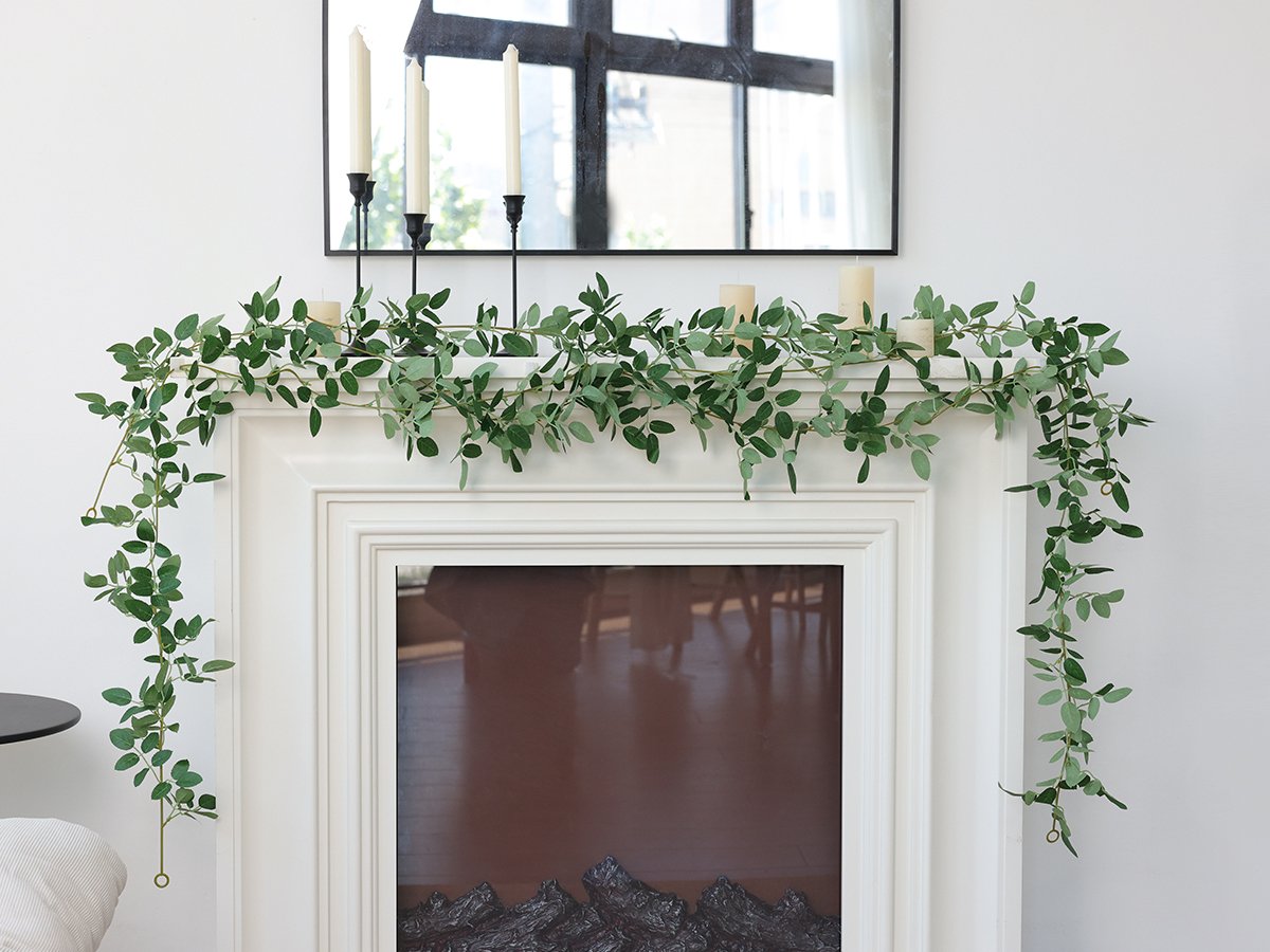 Artificial green vine garland draped over a white fireplace mantel with candles and a mirror above.