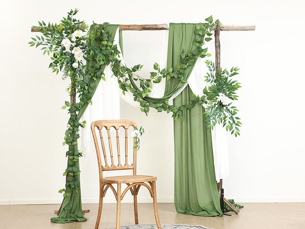 Wedding arch decorated with artificial green vines and white flowers, with draped green and white fabric and a wooden chair.