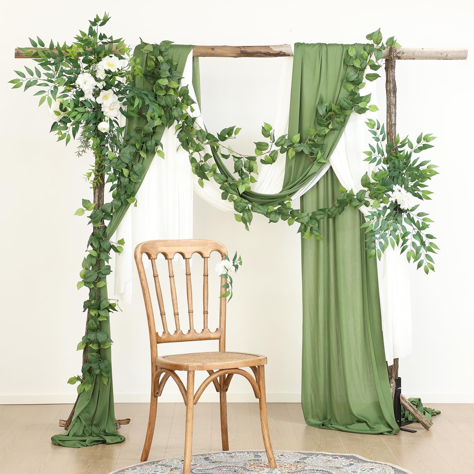Wedding arch decorated with artificial green vines and white flowers, with draped green and white fabric and a wooden chair.