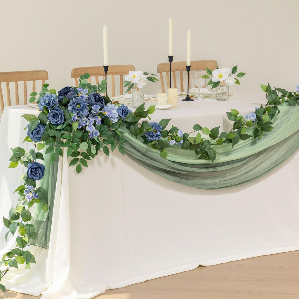 Artificial blue rose and greenery garland draped over a white tablecloth on a table with chairs and candles