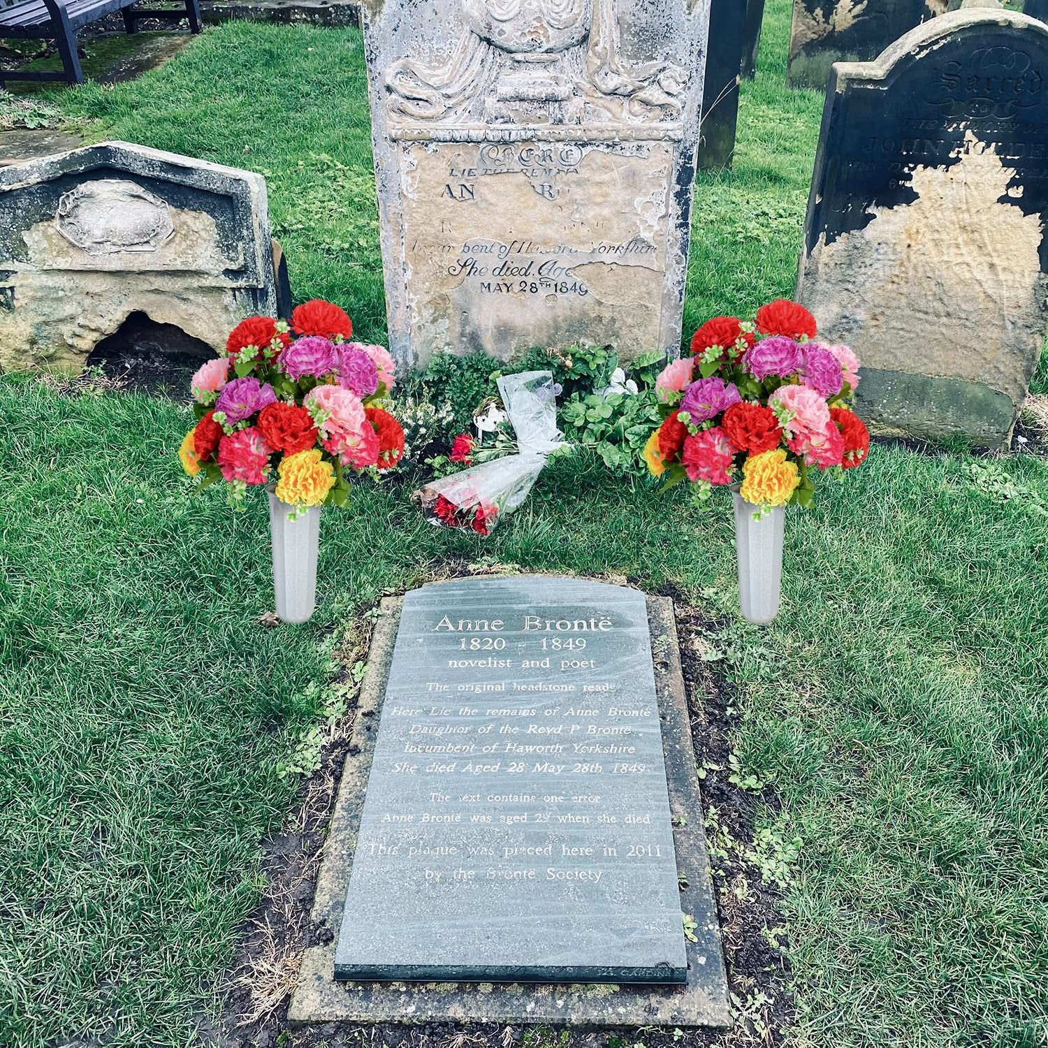 Two artificial carnation bouquets in vases placed at a cemetery headstone, with other gravestones in the background.