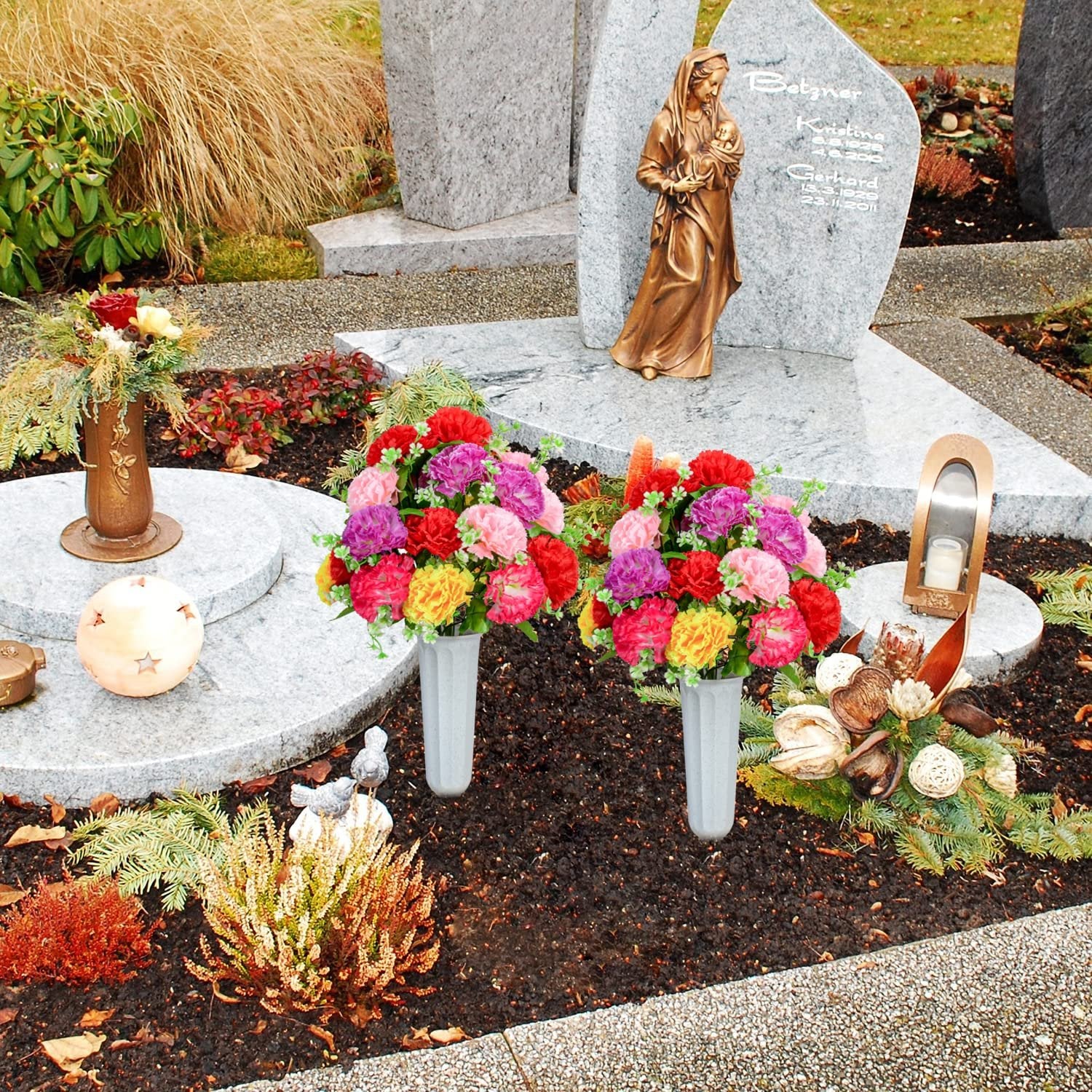 Two artificial carnation bouquets in vases placed at a cemetery grave with a statue and headstone.