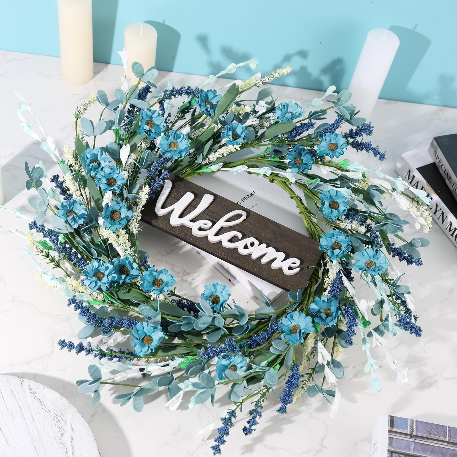 Artificial blue forsythia wreath with a wooden 'Welcome' sign, set against a light blue background and white candles.