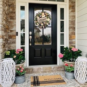 White magnolia wreath with buffalo plaid bow and 'BLESSED' plaque hanging on a black front door, flanked by potted flowers and lanterns.
