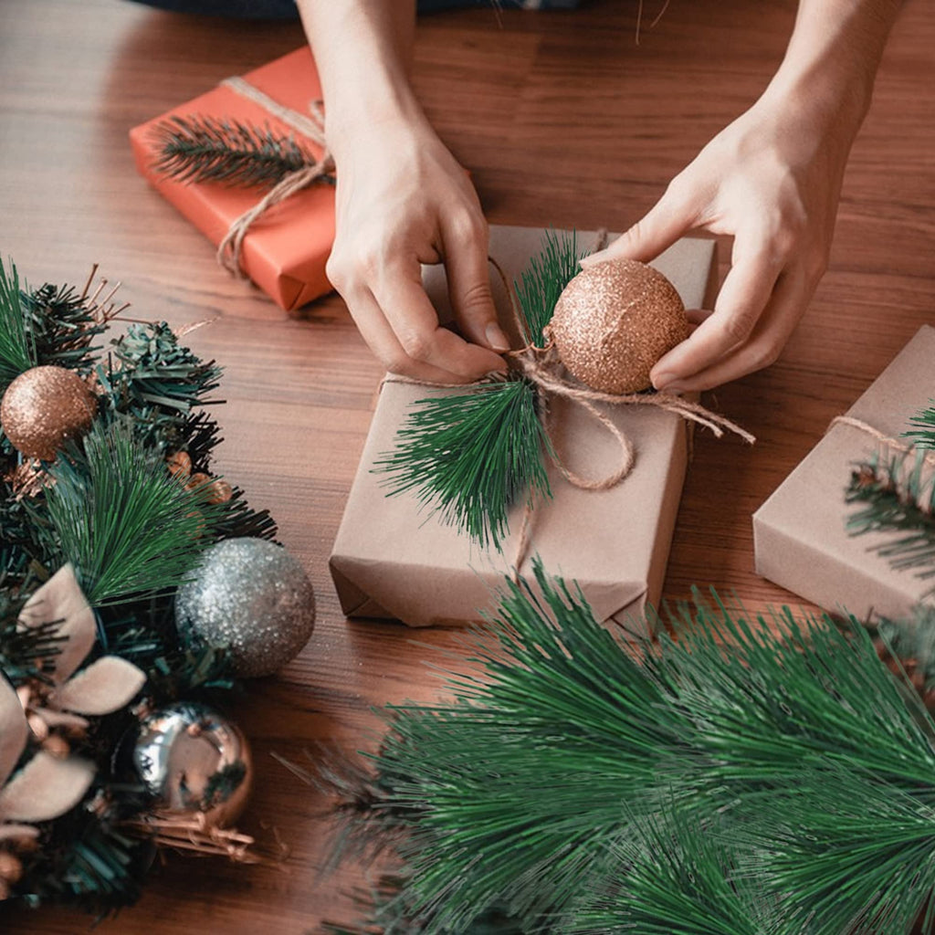 Hands decorating a gift box with pine sprigs and a gold ornament for the holidays.