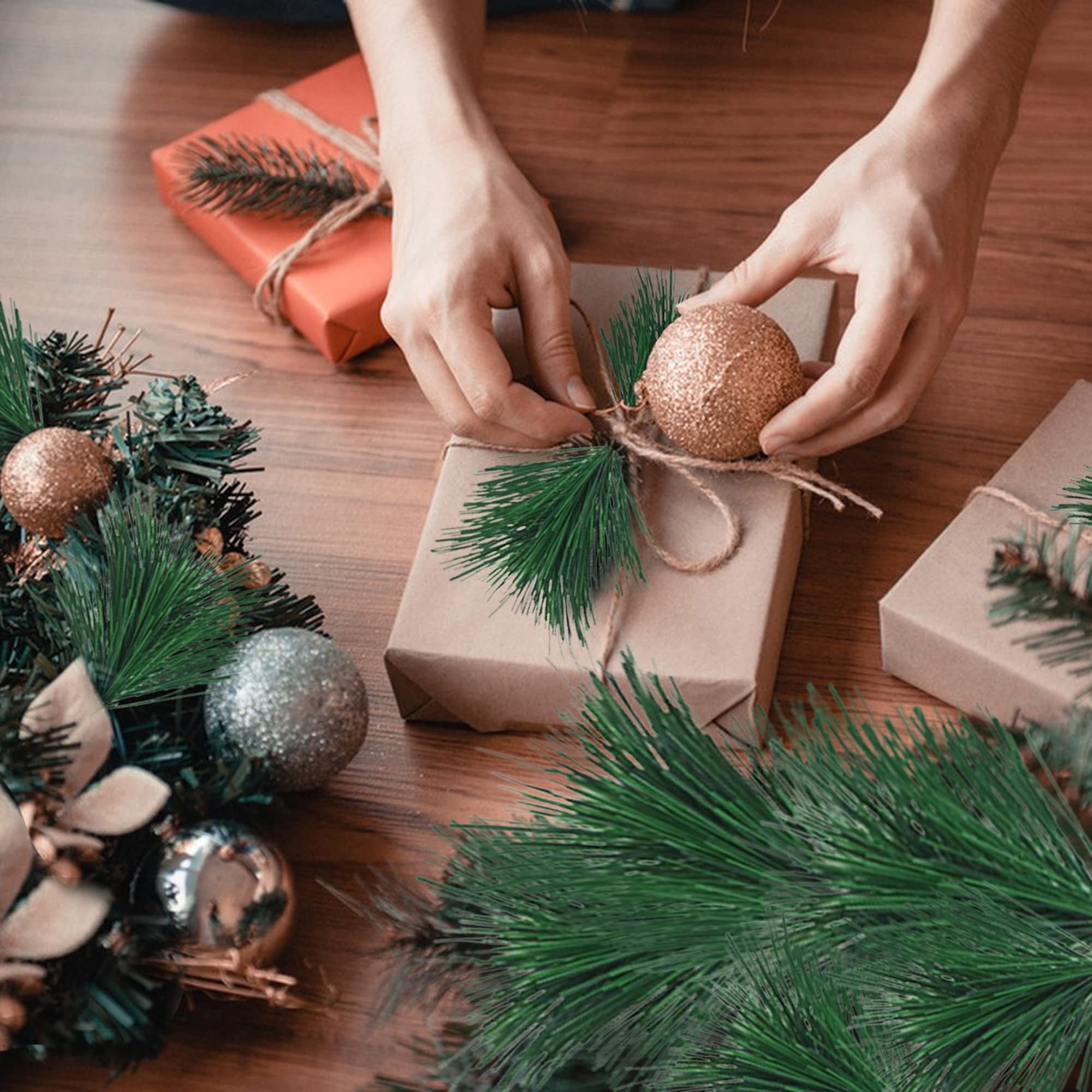 Hands decorating a gift box with pine sprigs and a gold ornament for the holidays.