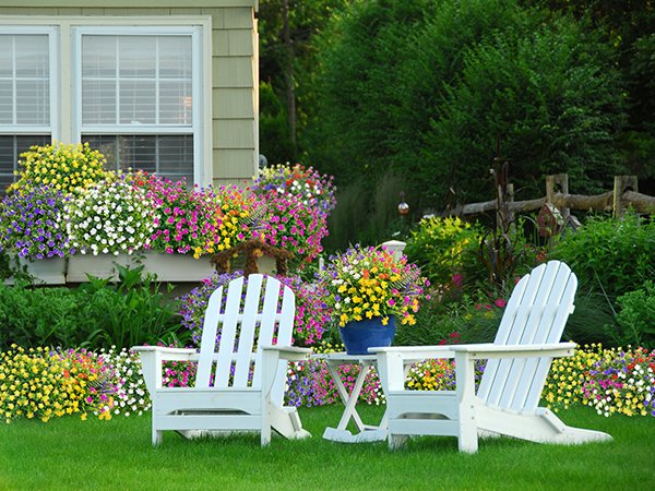 Two white Adirondack chairs on a green lawn in front of a house with colorful flowers in window boxes and pots.