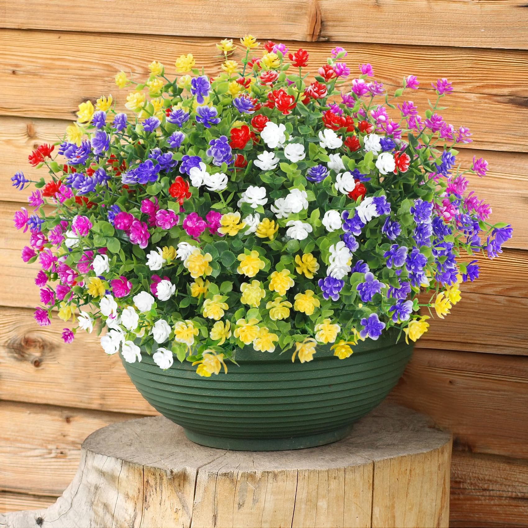 Artificial flowers in a green planter on a wooden stump against a wooden wall, featuring red, purple, white, yellow, and magenta blooms.