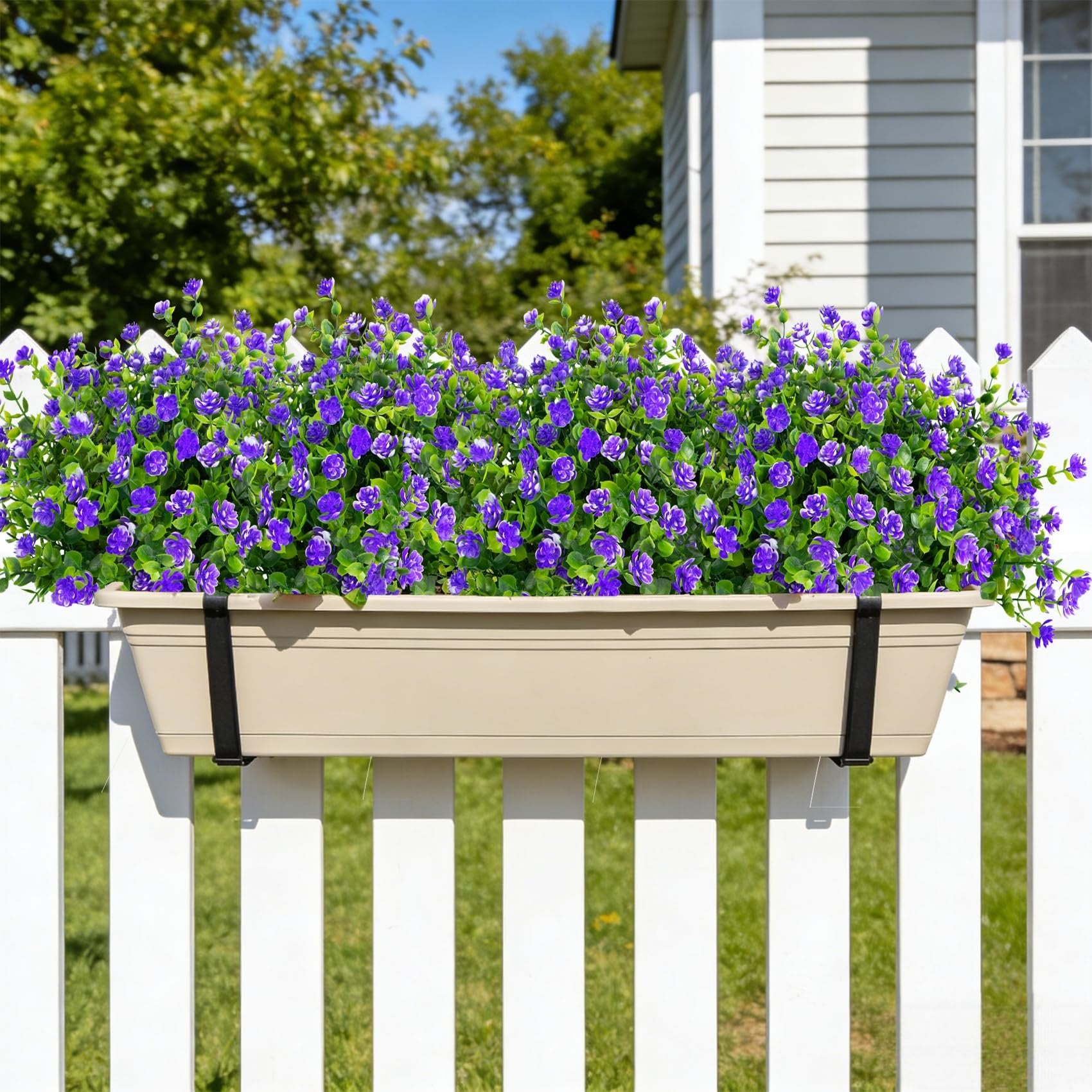 Tan planter box filled with purple artificial flowers attached to a white picket fence