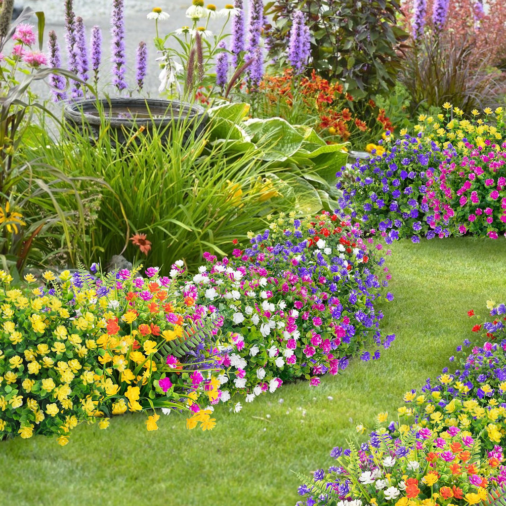 Artificial flowers in red, purple, magenta, white, and yellow arranged in a garden setting with green grass and other plants.
