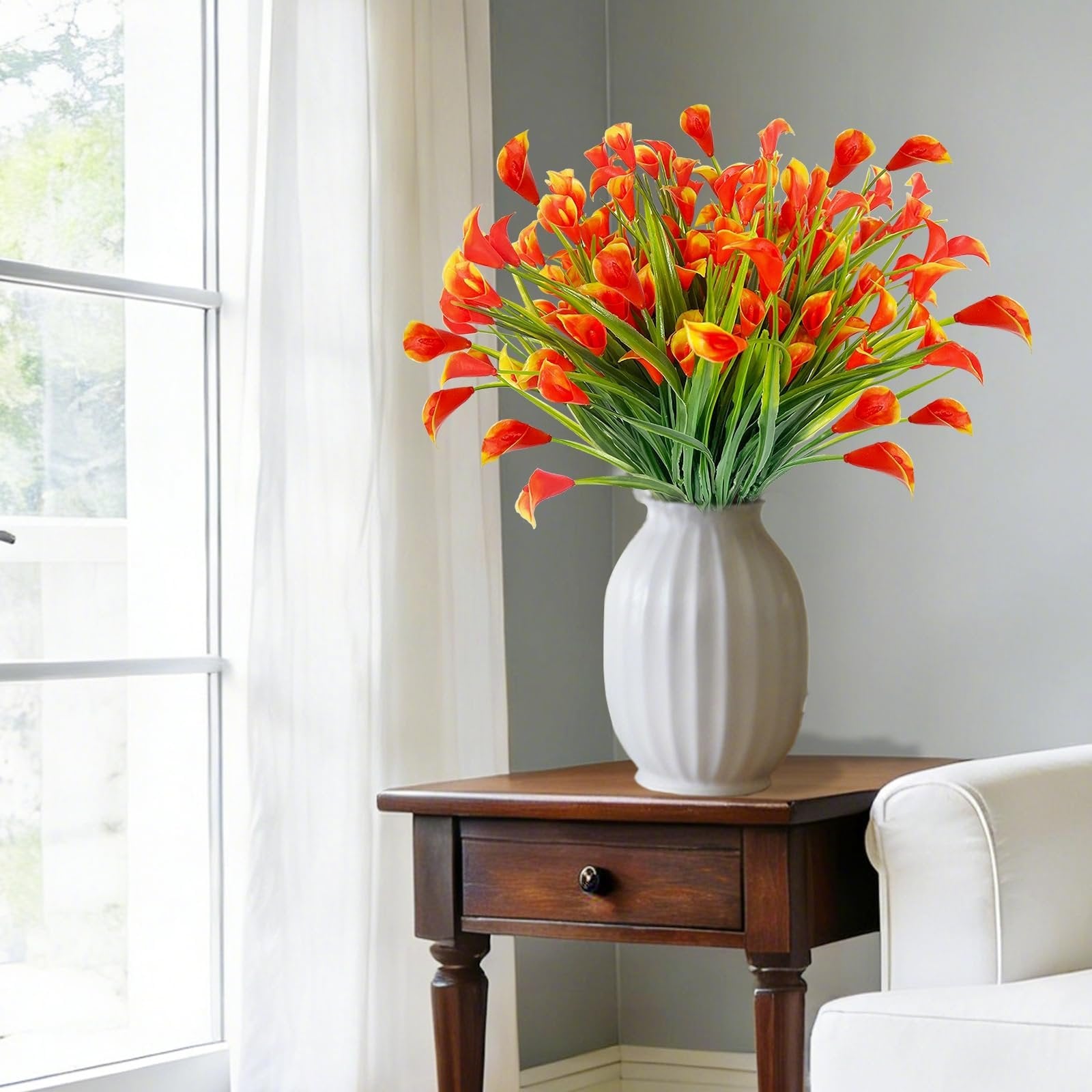 Bouquet of orange and yellow artificial calla lilies in a white vase on a wooden side table.