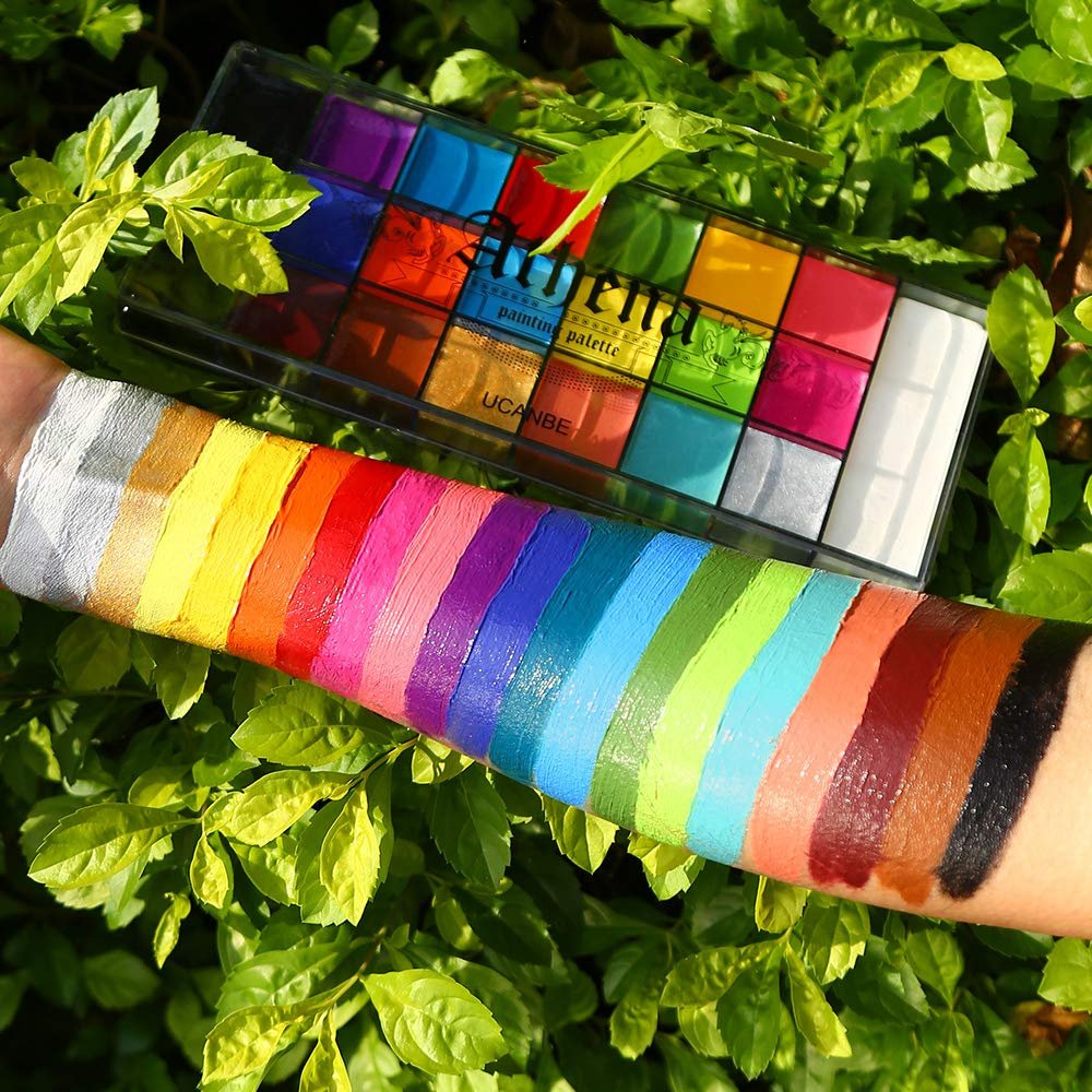 Arm with swatches of 20 oil-based face and body paints next to an open palette of the same colors, surrounded by green leaves.