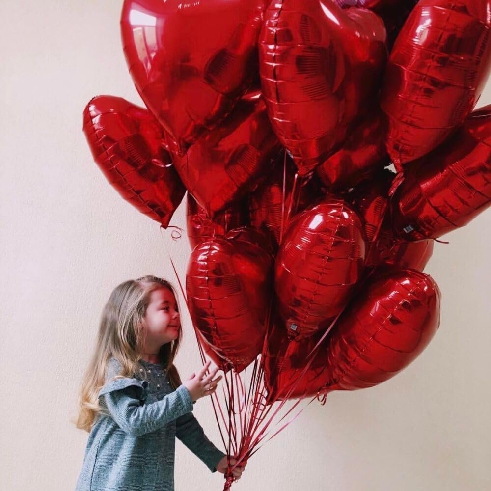 Young girl holding a large bunch of red heart-shaped foil balloons against a plain background.