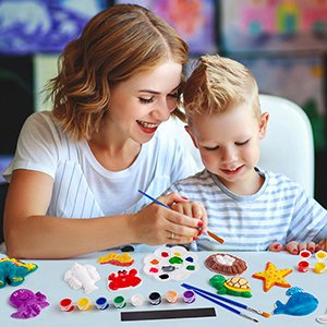 Mother and son painting plaster figurines together at a table with paints and brushes