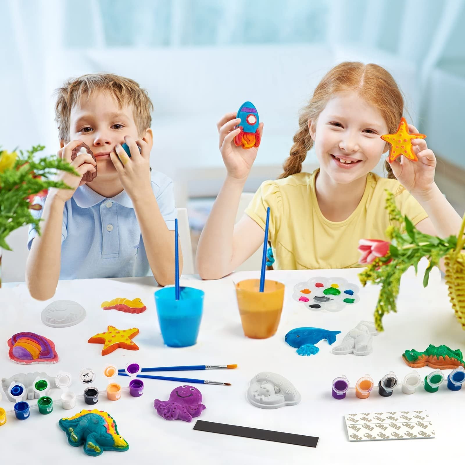Two children happily painting plaster figurines including a rocket and a starfish, with paints and brushes laid out on a table.