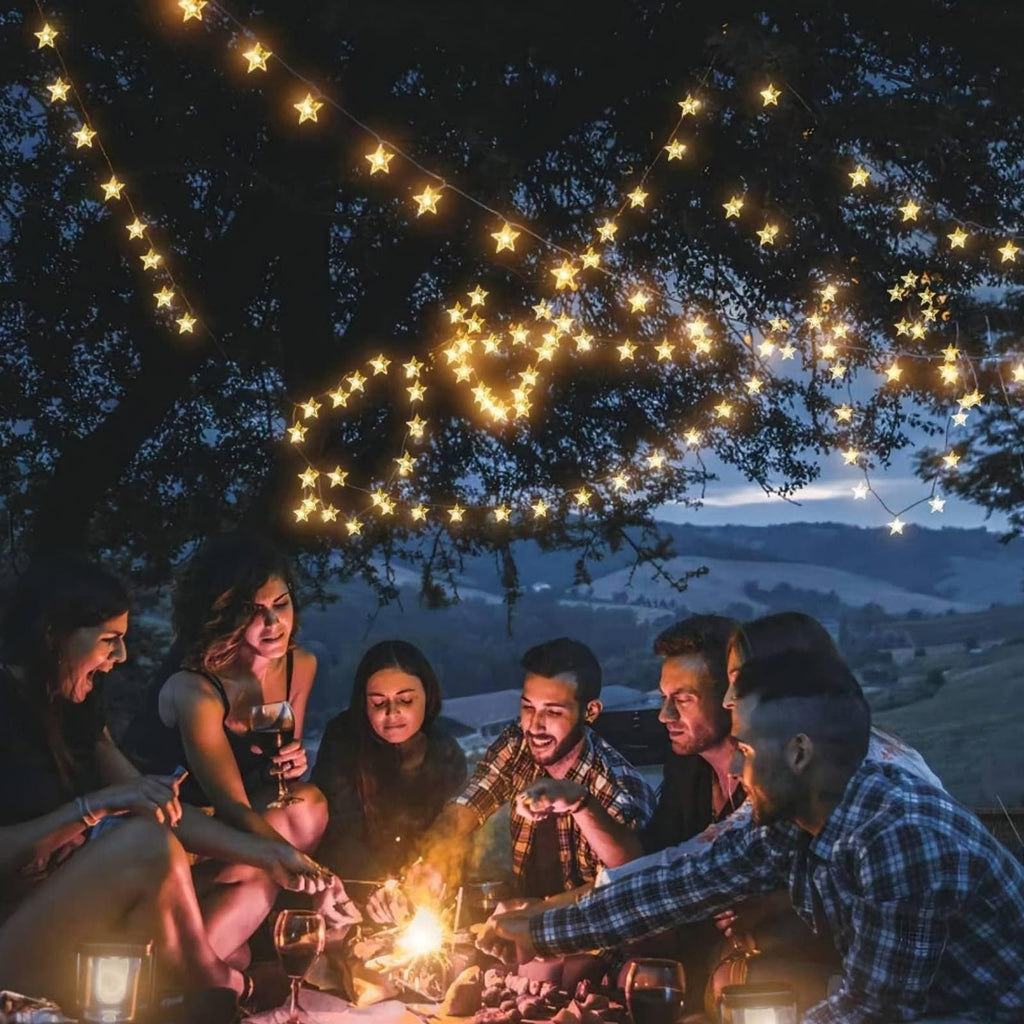 Group of friends gathered around a campfire at night, with warm white star-shaped string lights overhead.
