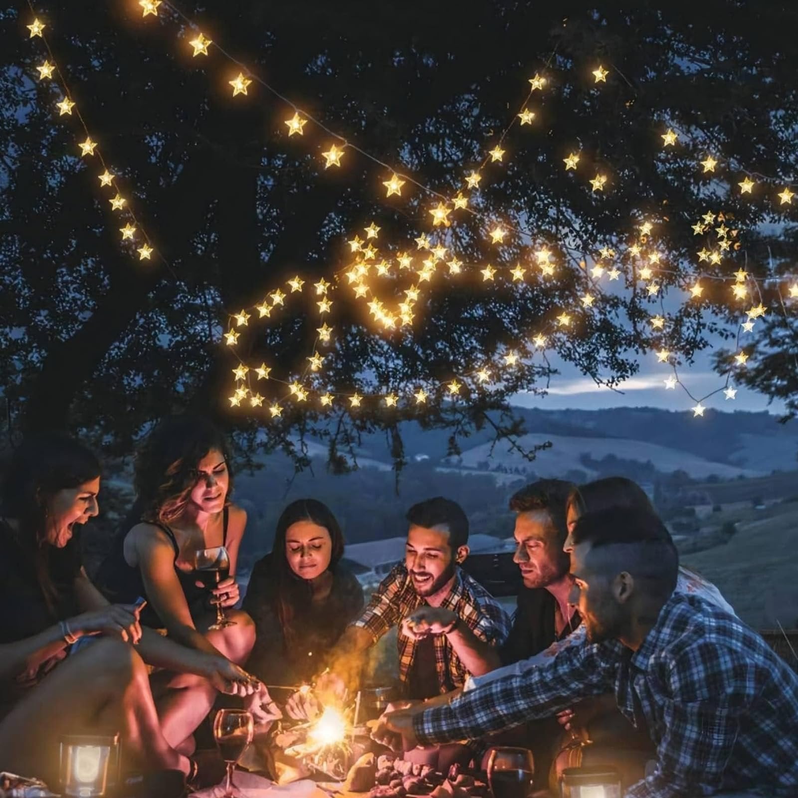 Group of friends gathered around a campfire at night, with warm white star-shaped string lights overhead.