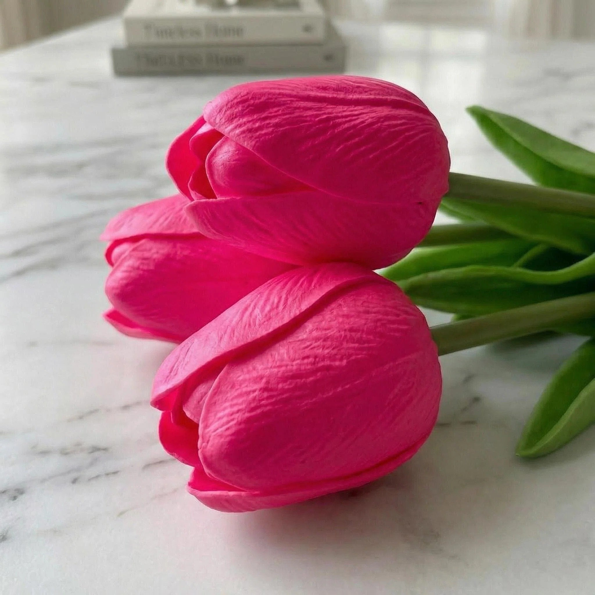 Close-up of three hot pink artificial tulip flower heads with green stems and leaves on a marble surface, with blurred books in the background.