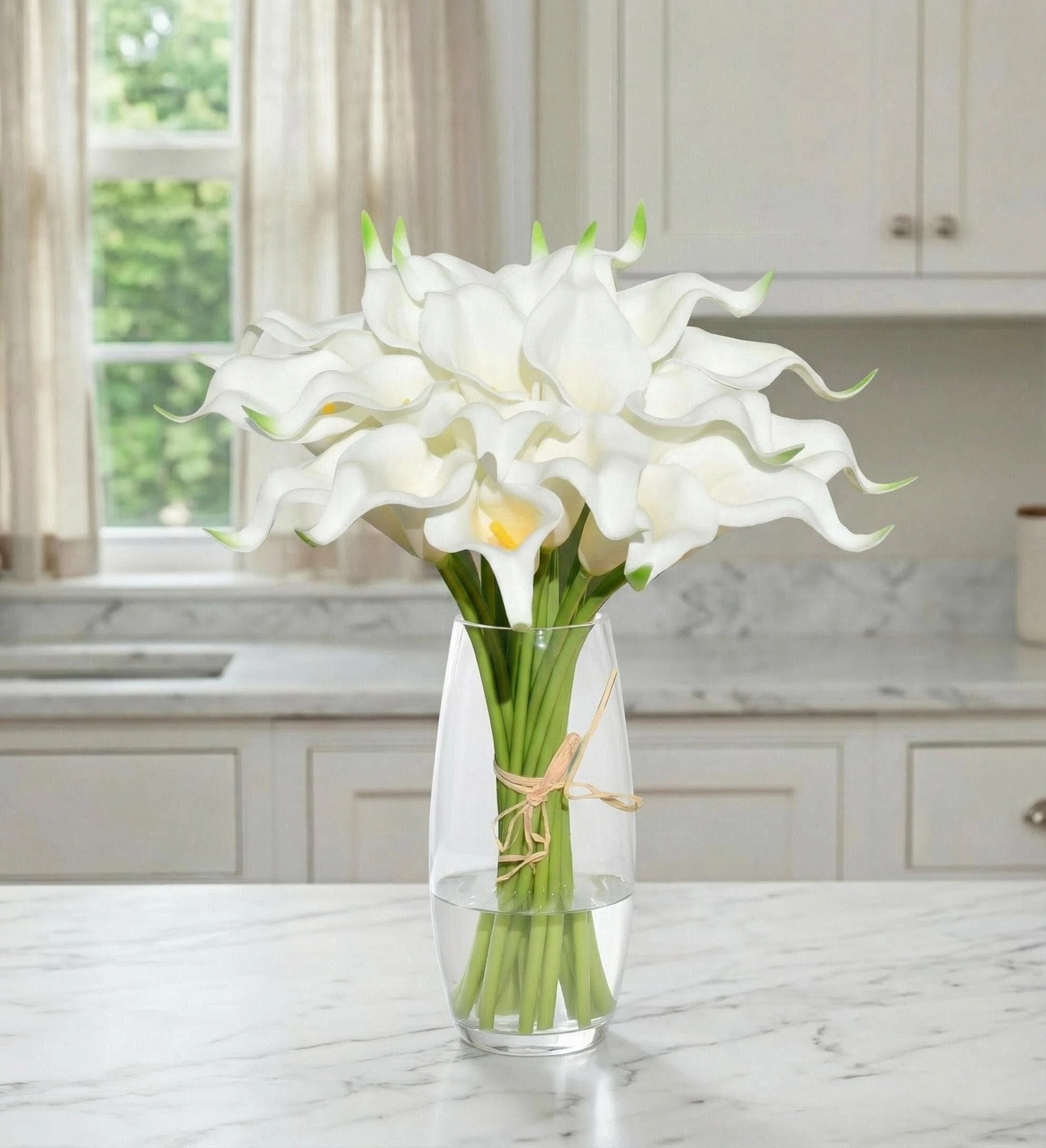 Bouquet of white calla lilies in a glass vase on a marble countertop with kitchen cabinets and window in background