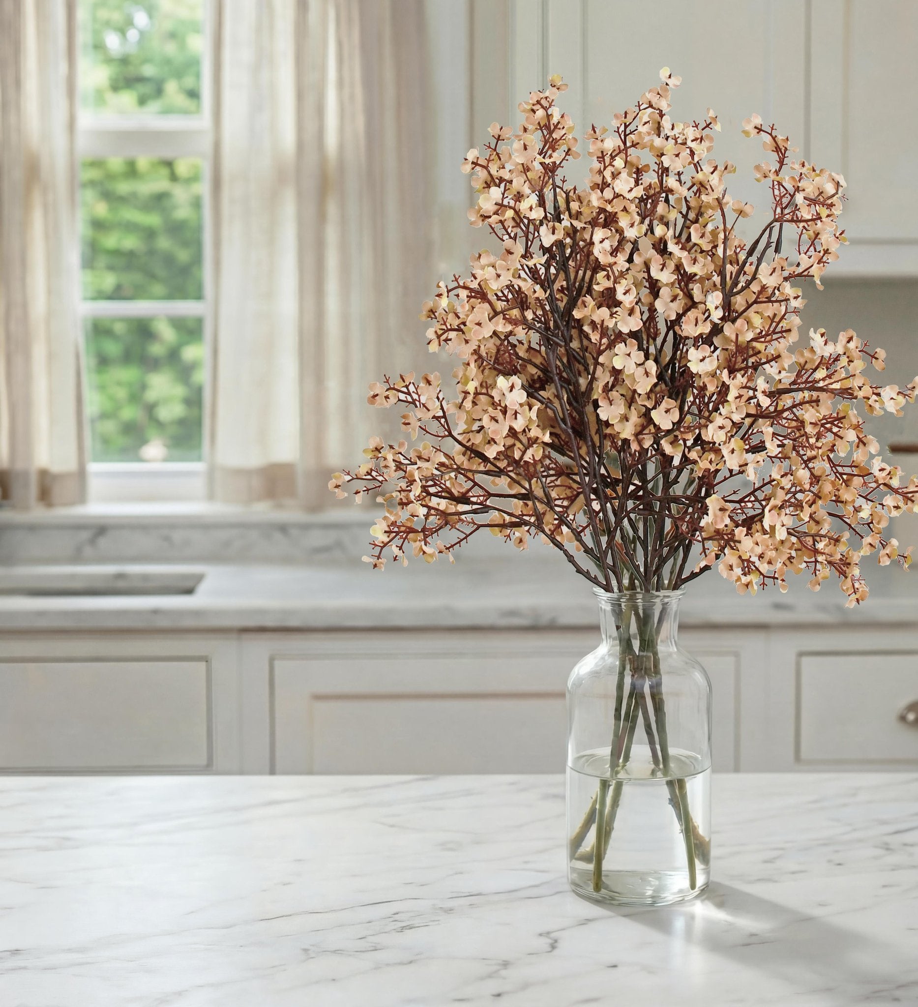 Bouquet of light peach artificial flowers in a clear glass vase on a marble countertop, with a window and curtains in the background.