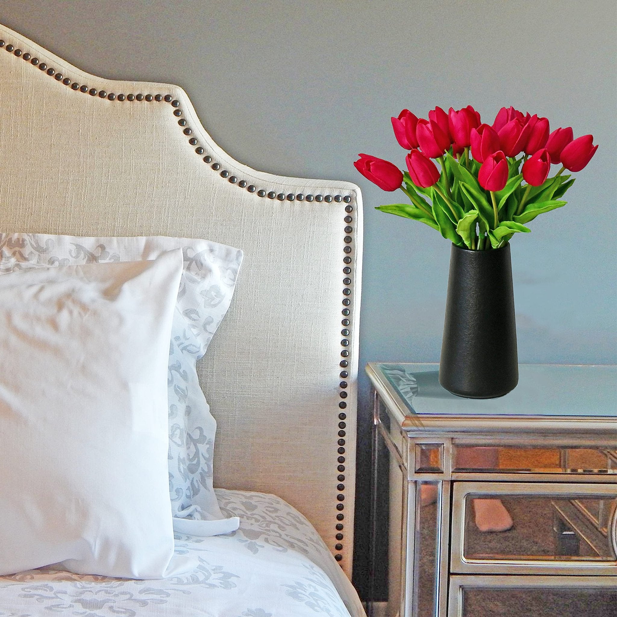 A vase of hot pink artificial tulips sits on a mirrored nightstand next to a tufted headboard.