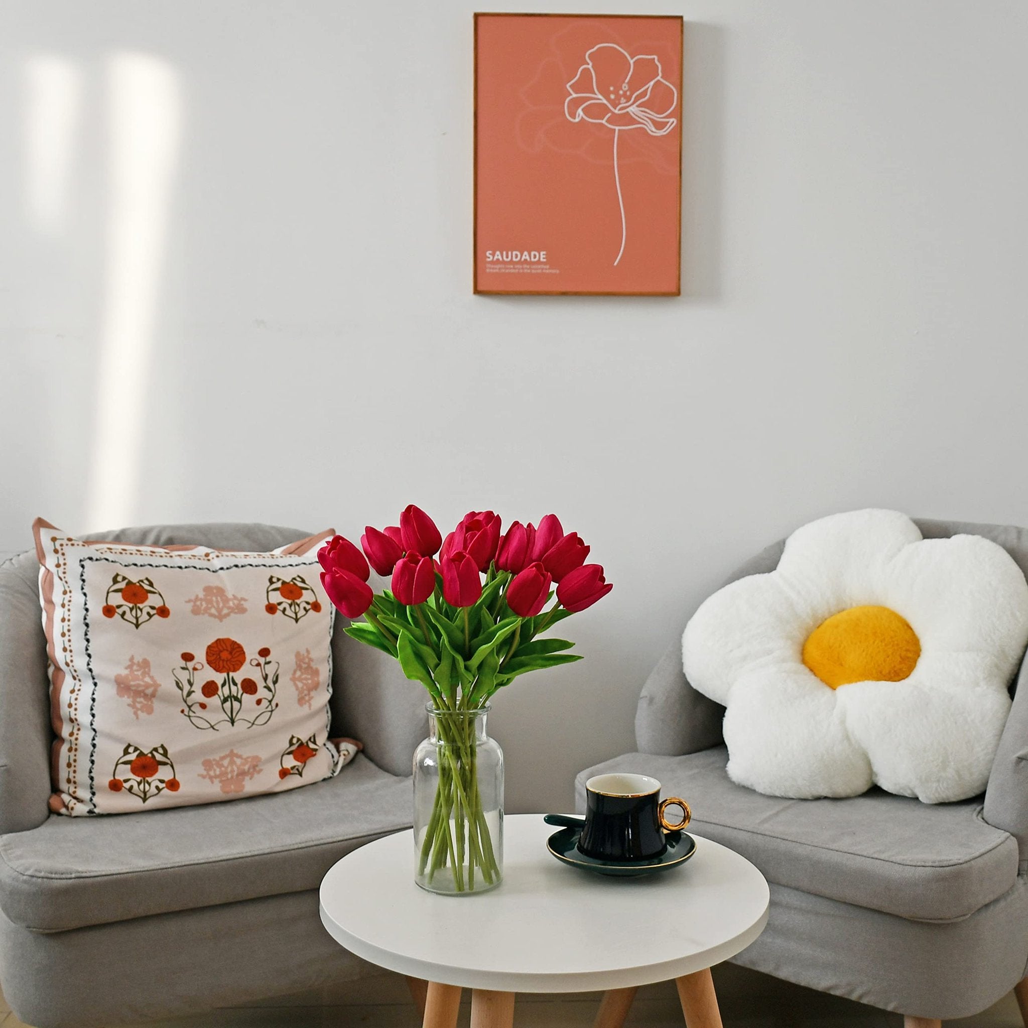 A vase of hot pink artificial tulips sits on a white round table between two gray chairs, with a framed picture on the wall above.