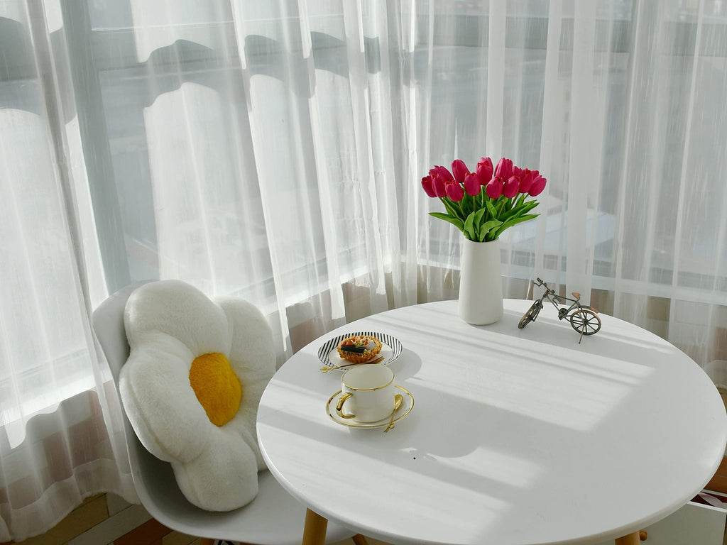 A bouquet of hot pink artificial tulips in a white vase on a round white table with a pastry and teacup, next to a chair with a flower pillow.