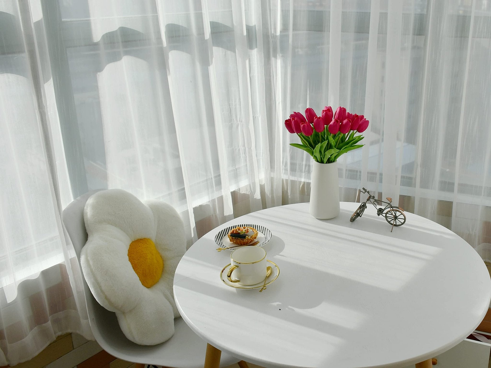 A bouquet of hot pink artificial tulips in a white vase on a round white table with a pastry and teacup, next to a chair with a flower pillow.