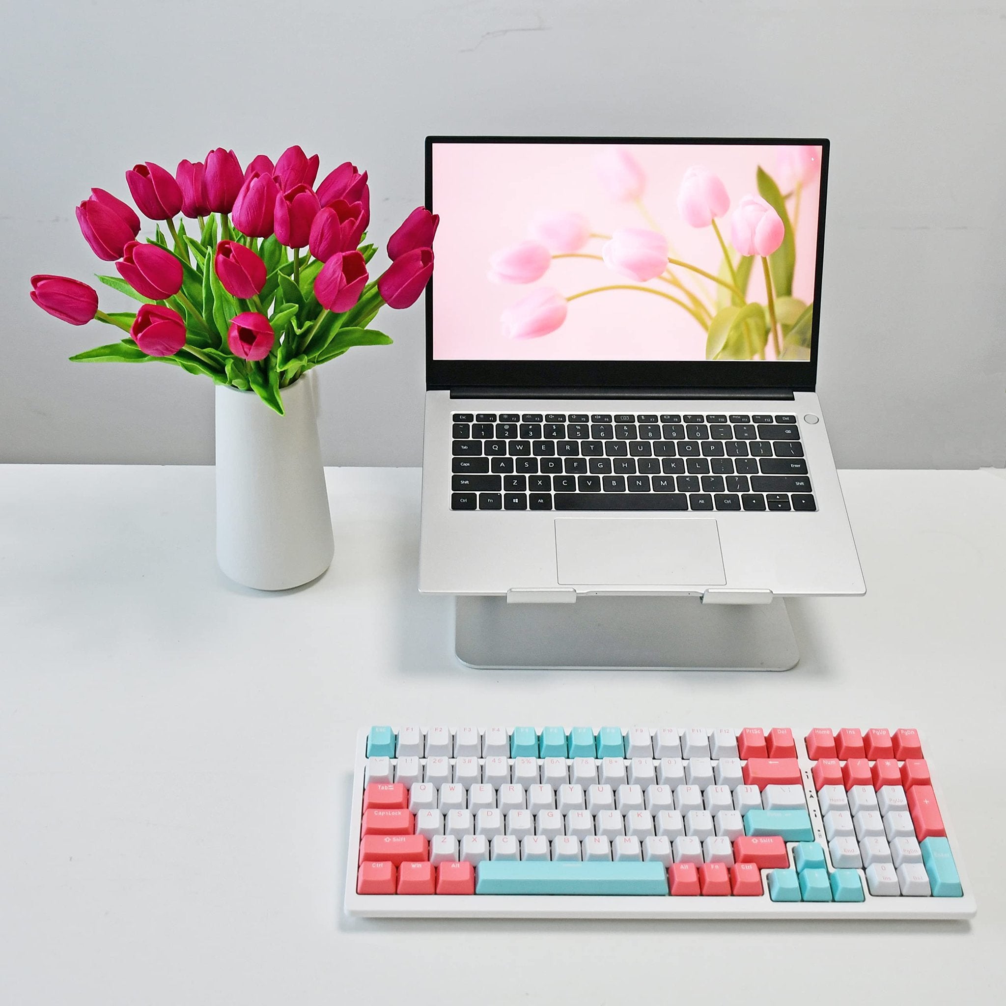 A vase of hot pink artificial tulips sits next to a laptop displaying pink tulips, with a colorful keyboard in the foreground.