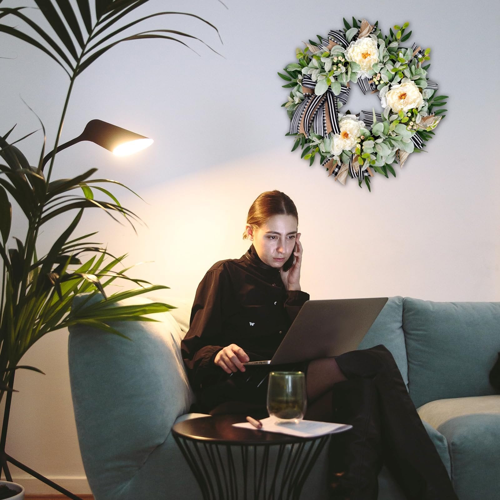 Woman on couch with laptop and phone next to a decorative wreath on the wall