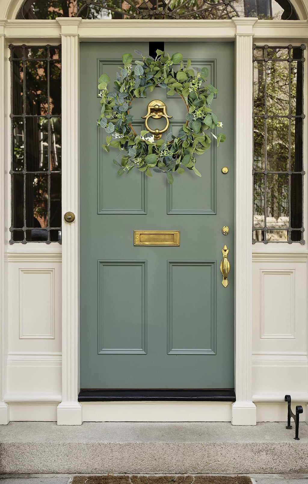 Artificial eucalyptus wreath with silk leaves and white flowers hanging on a green front door with brass hardware.