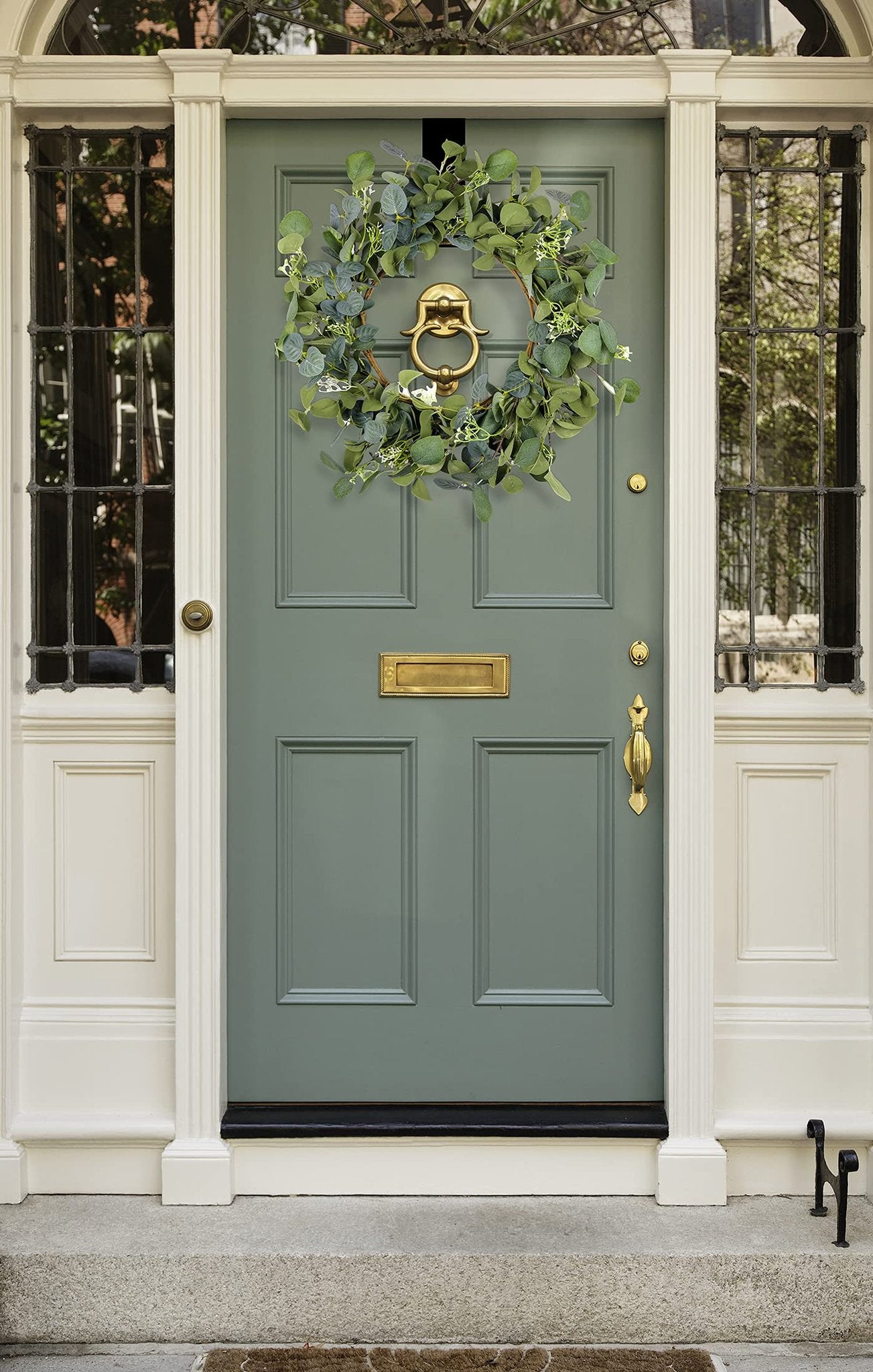 Artificial eucalyptus wreath with silk leaves and white flowers hanging on a green front door with brass hardware.