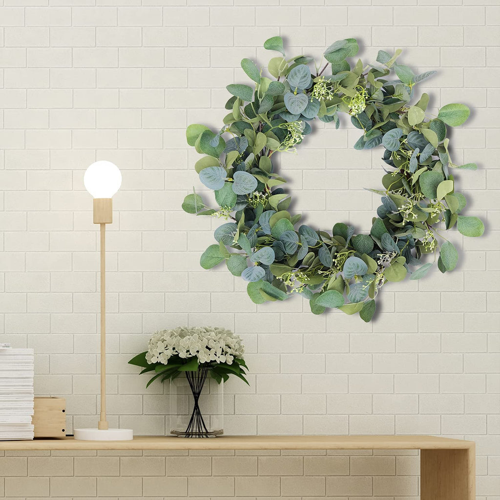 Artificial eucalyptus wreath with silk leaves hanging on a white brick wall above a wooden shelf with a lamp and flowers.