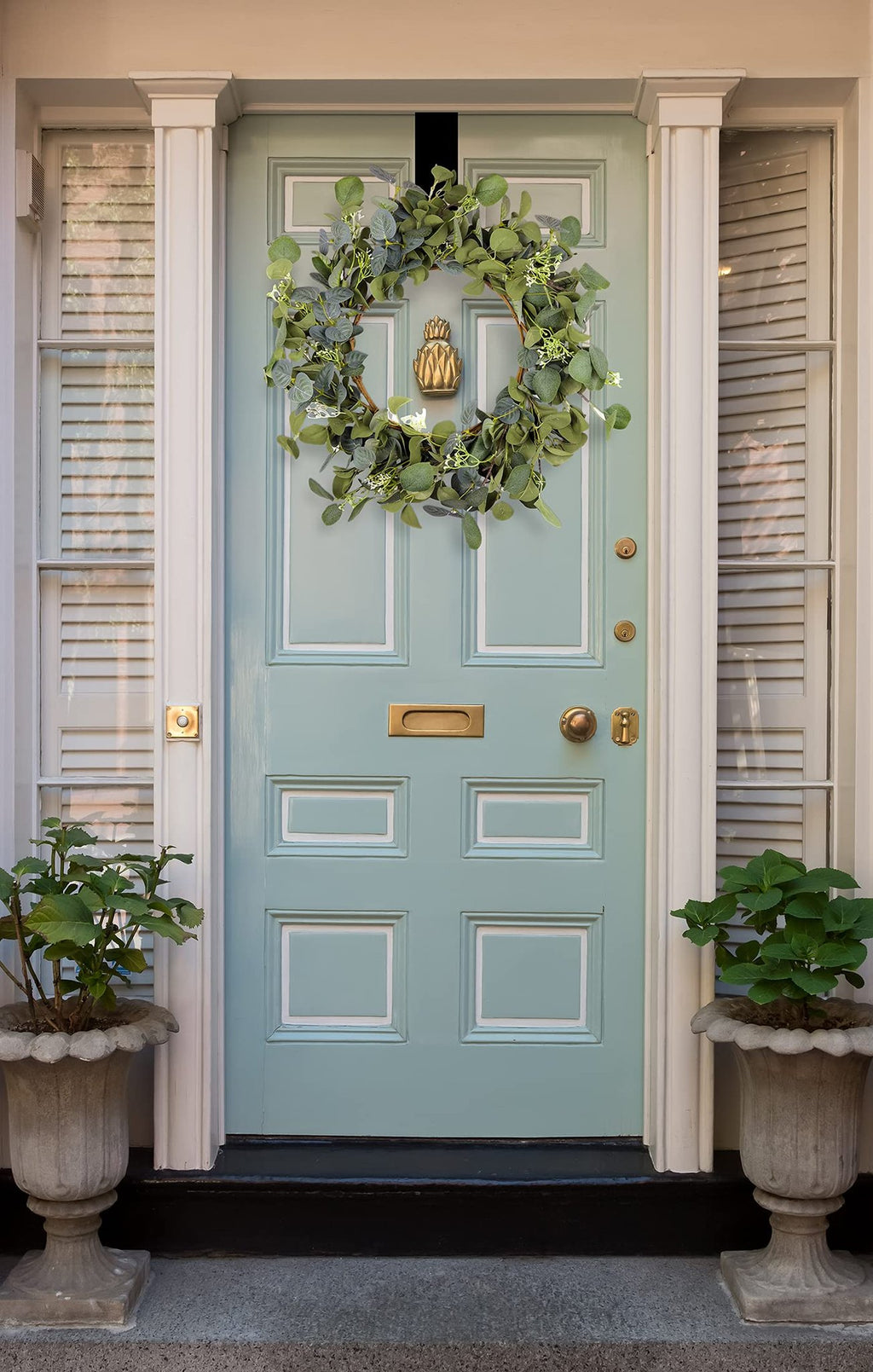 Artificial eucalyptus wreath with silk leaves hanging on a light blue front door with brass hardware and planters.