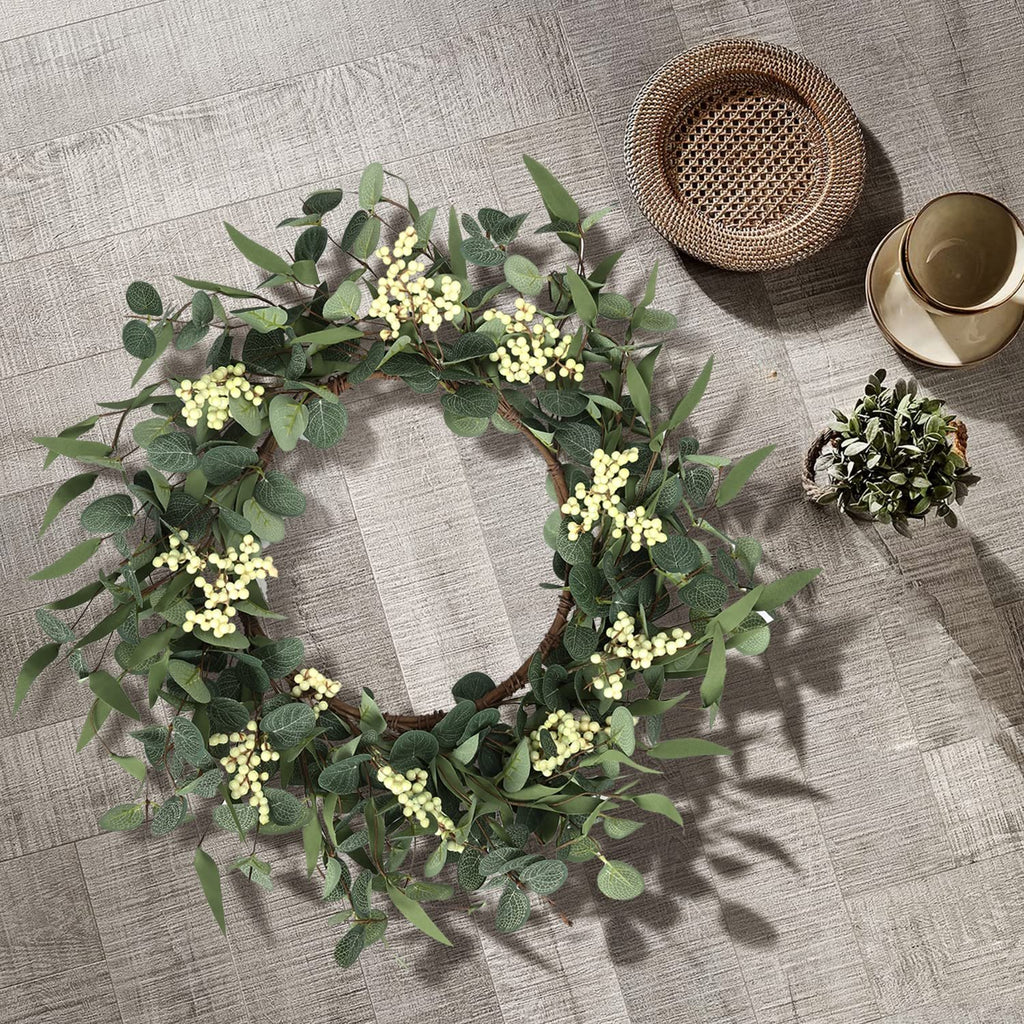 Eucalyptus wreath with white berries arranged on a wooden floor next to a small plant and a cup and saucer.