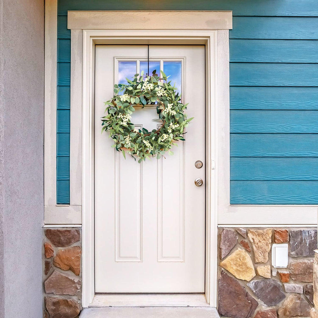 Eucalyptus wreath with white berries hanging on a white front door with blue siding and stone accents.