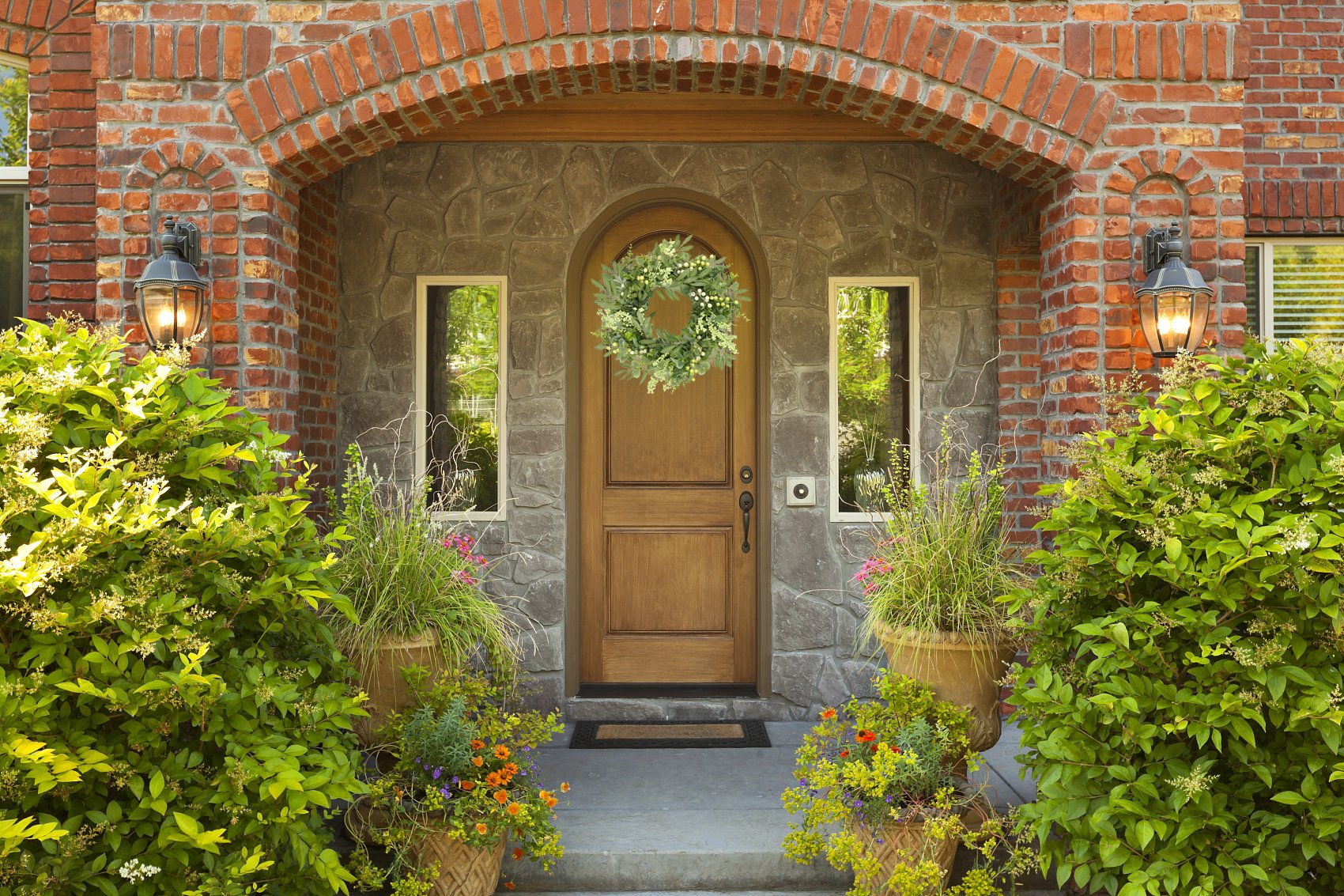 Eucalyptus wreath with olive leaves and berries hanging on a wooden front door, flanked by windows and potted plants.