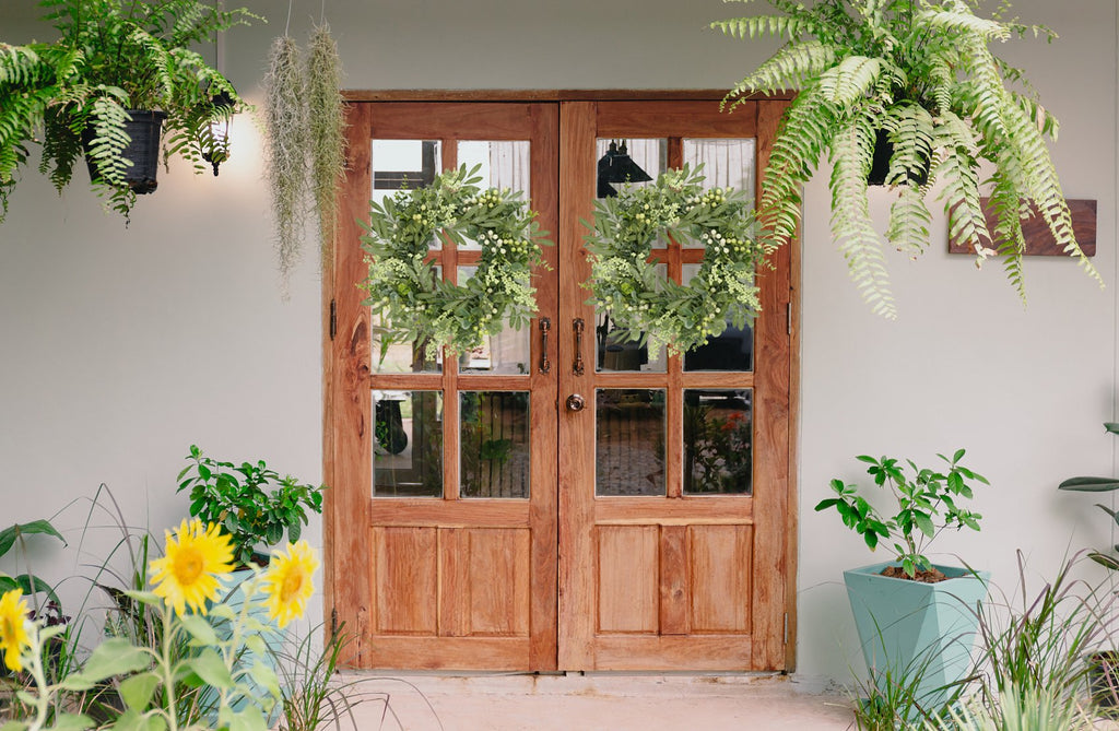 Two eucalyptus wreaths with olive leaves and berries hanging on a wooden double door, flanked by plants.