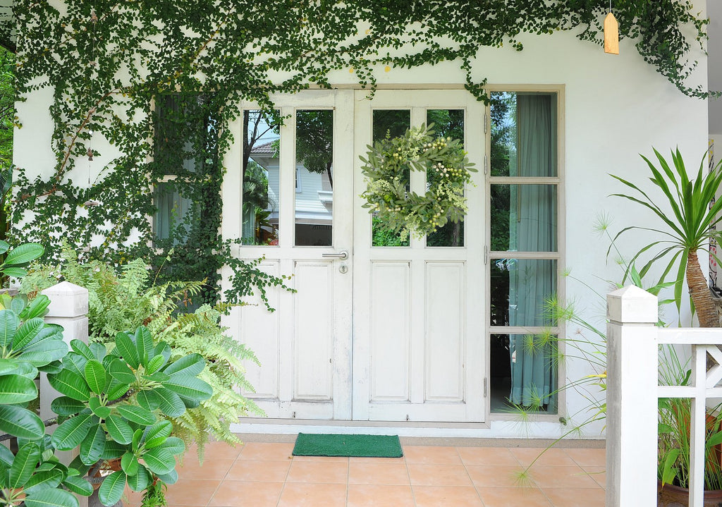 Front door with a green wreath adorned with olive leaves and berries, surrounded by lush greenery and plants.