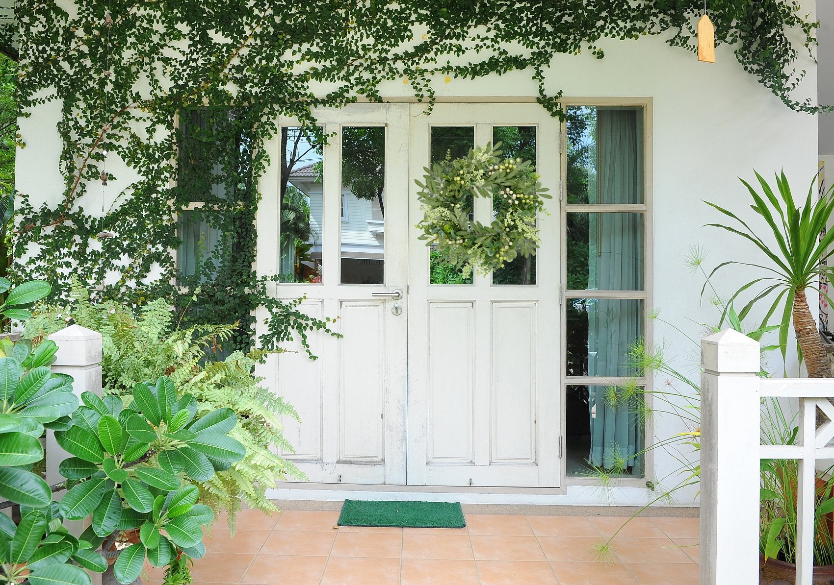 Front door with a green wreath adorned with olive leaves and berries, surrounded by lush greenery and plants.